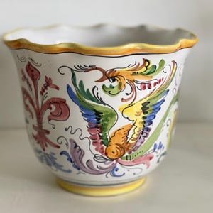 Könnte beinhalten: Ein weißer Keramik-Blumentopf mit gewelltem Rand und gelbem Sockel. Der Topf ist mit farbenfrohen, handgemalten Designs verziert, darunter ein Vogel und florale Motive. Der Vogel hat gelbe, grüne, blaue und orangefarbene Federn.