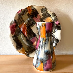 Può includere: Un set decorativo composto da vaso e piatto in ceramica. Il vaso presenta una parte superiore svasata e una base ampia, con un motivo di pennellate arancioni, viola e grigie. Il piatto ha un bordo ondulato e un design astratto simile.