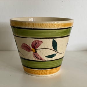 Könnte beinhalten: Ein cremefarbener Keramik-Blumentopf mit floralem Design. Der Topf hat eine strukturierte Oberfläche mit gelben, grünen und schwarzen Bändern. Auf der Vorderseite ist eine rosa und gelbe Blume mit grünen Blättern aufgemalt. Geeignet für den Innen- und Außenbereich.