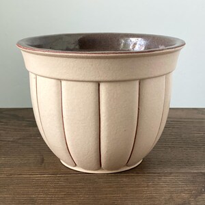 Può includere: Un vaso di ceramica beige con dettagli a coste verticali. Il bordo e l'interno sono di un marrone più scuro. Il vaso poggia su una superficie di legno. Questo pezzo decorativo è adatto per uso interno o esterno.