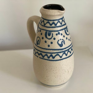 Può includere: Un'anfora in ceramica bianca con un motivo geometrico blu e bianco. L'anfora ha un corpo arrotondato e un beccuccio.