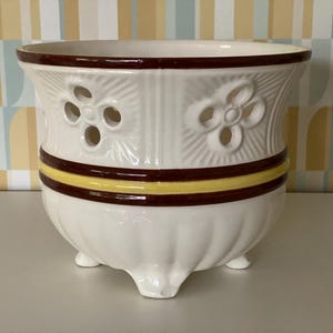 Op de afbeelding: Witte keramische bloempot met een decoratief bloemmotief en drie kleine voetjes. De pot heeft een donkerbruine rand en een gele en bruine band in het midden. De pot heeft uitgesneden bloemvormen.