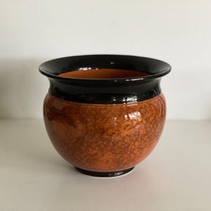 Op de afbeelding: Decoratieve keramische bloempot met een glanzende afwerking. Zwarte rand en basis, een getextureerde oranje-bruine body. De binnenkant is lichter oranje. Geschikt voor binnen of buiten. Diameter ongeveer 20 cm.