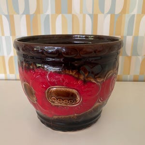 Può includere: Un vaso di ceramica con finitura lucida. Il vaso presenta un bordo e una base marrone scuro, con un vivace motivo floreale rosso sul davanti. Il design include accenti ovali marroni. Un pezzo decorativo per piante.