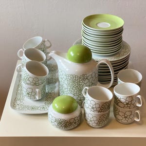 Könnte beinhalten: Ein weißes Keramik-Teeservice mit grünem Blumenmuster. Das Set umfasst eine Teekanne, eine Zuckerdose, Tassen, Untertassen und ein Serviertablett. Die Teekanne und die Zuckerdose haben grüne Deckel.
