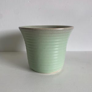 Op de afbeelding: Een lichtgroene keramische vaas met een geribbelde textuur. De vaas heeft een licht uitlopende rand met een lichtbruine rand. De vaas staat op een wit oppervlak.