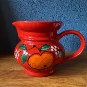 Könnte beinhalten: Eine rote Keramikkanne mit einem weißen und grünen Apfeldesign. Die Kanne hat einen Henkel und eine breite Öffnung.