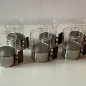 Könnte beinhalten: Satz mit sechs klaren Glasbechern mit Edelstahlsockeln und Holzgriffen. Die Becher haben ein dekoratives Muster am Rand.