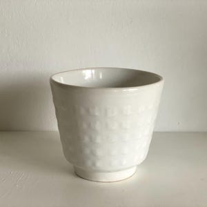 Könnte beinhalten: Eine weiße Keramiktasse mit einem strukturierten, sich wiederholenden Muster. Die Tasse hat eine abgerundete Form mit einem leicht ausgestellten Rand und einem kleinen, kreisförmigen Boden. Die Tasse ist etwa 10 cm hoch.