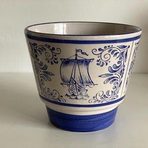 Può includere: Un vaso di ceramica con un design nautico blu e bianco. Il vaso presenta una barca a vela blu e decorazioni a volute su uno sfondo color crema. La base e il bordo sono dipinti di blu.