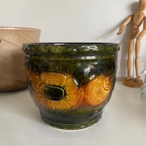 Può includere: Un vaso di ceramica con un motivo floreale verde scuro e arancione. Il vaso ha una forma arrotondata con una superficie strutturata e un bordo decorativo. La tavolozza dei colori comprende sfumature di verde, arancione e marrone, creando un'estetica vintage.