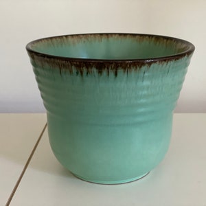 Könnte beinhalten: Ein hellgrüner Keramiktopf mit braunem Rand. Der Topf hat einen leicht ausgestellten Rand und einen abgerundeten Boden.