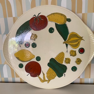 Könnte beinhalten: Eine runde, cremefarbene Servierplatte mit einem farbenfrohen Gemüse-Design. Die Illustration zeigt eine rote Tomate, eine gelbe Zitrone, eine grüne Gurke und Mais. Die Platte hat zwei Griffe und einen geriffelten Rand.