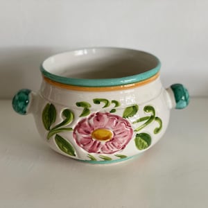 Könnte beinhalten: Weißer Keramiknapf mit floralem Design. Die Schale hat eine rosa Blume mit gelbem Zentrum, grüne Blätter und türkisfarbene und orangefarbene Verzierungen. Sie hat zwei türkisfarbene Griffe auf beiden Seiten.