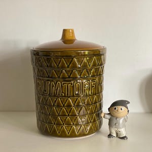 Op de afbeelding: Een vintage keramische pot met een deksel, met een geometrisch patroon en het woord "RUMTOPF" in reliëf op de voorkant. De pot is donker olijfgroen met een bruin deksel. Een klein beeldje van een kabouter staat naast de pot.