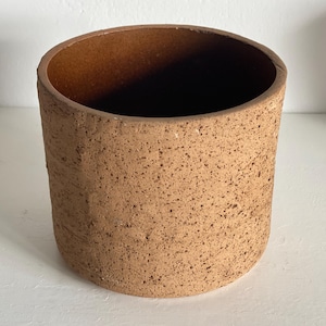 Op de afbeelding: Een cilindrische, handgemaakte keramische pot met een gespikkelde, getextureerde oppervlakte. De pot heeft een warme, aardse bruine kleur en een gladde binnenkant. De open bovenkant onthult een donkere binnenkant. Deze decoratieve pot is geschikt voor planten of opslag.