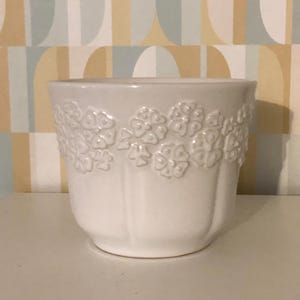 Op de afbeelding: Een witte keramische bloempot met een bloemenreliëf aan de bovenkant. De pot heeft een afgeronde vorm en staat op een wit oppervlak. De achtergrond is een behang met een geometrisch patroon.