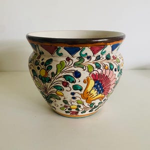 Può includere: Un vaso di fiori in ceramica con base color crema, ornato da motivi floreali colorati. Il bordo è marrone e il design comprende elementi blu, rosa, gialli e verdi. Il vaso ha una forma ampia e arrotondata.