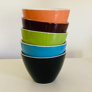 Op de afbeelding: Een stapel van vijf kleurrijke keramische kommen. De kommen zijn in elkaar genest, beginnend met een zwarte kom onderaan, gevolgd door een blauwe kom, een groene kom, een bruine kom en een oranje kom bovenaan.