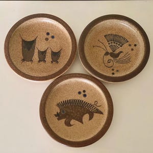 Könnte beinhalten: Drei runde Keramikplatten mit braunen Rändern und gesprenkelten, beigefarbenen Zentren. Jede Platte zeigt ein anderes Tiermotiv in Braun und Schwarz: Eulen, einen Vogel und einen Igel. Ein dekoratives Set.