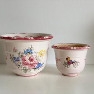 Pode incluir: Duas floreiras de cerâmica. A floreira maior apresenta um desenho floral com rosas cor-de-rosa e flores azuis. A floreira menor tem um desenho de pássaro. Ambas as floreiras têm uma base branca com uma borda vermelha.