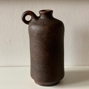Könnte beinhalten: Eine dunkelbraune Keramikvase mit Henkel. Die Vase hat eine strukturierte Oberfläche und eine abgerundete Form.