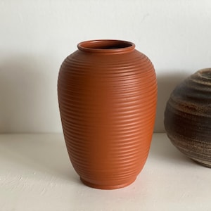 Op de afbeelding: Een terracotta-kleurige keramische vaas met een geribbelde textuur. De vaas heeft een afgeronde vorm met een smalle hals en een brede basis. Een tweede, kleinere, donkerbruine vaas is gedeeltelijk rechts zichtbaar. Woondecoratie.
