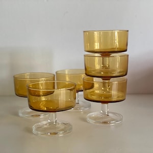 Könnte beinhalten: Ein Set aus sechs Dessertschalen aus bernsteinfarbenem Glas. Die Schalen haben eine abgerundete Form und stehen auf klaren, stielartigen Basen. Fünf Schalen sind auf einer weißen Oberfläche angeordnet, eine ist auf einem Vierer-Set gestapelt.