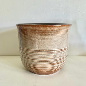 Può includere: Un vaso cilindrico in ceramica caratterizzato da una smaltatura bicolore con un bordo marrone scuro che sfuma in un corpo beige più chiaro. La superficie esterna è decorata con una serie di scanalature orizzontali incise lungo tutto il vaso.