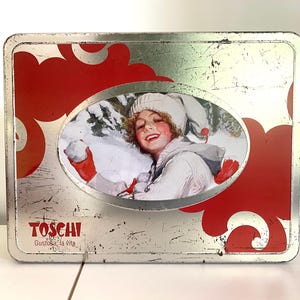 Può includere: Una scatola di latta vintage argentata con accenti rossi e bianchi. La scatola presenta una donna con un cappello e un cappotto bianchi che tiene una palla di neve. Il testo "TOSCANI" e "Gustosa la Vita" è stampato sulla scatola.