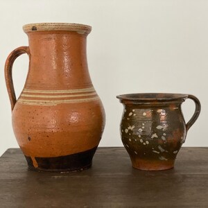 Può includere: Due brocche in terracotta marrone con smalto bianco e marrone. La brocca più grande ha un manico e un'ampia apertura. La brocca più piccola ha un manico e una forma più rotonda.