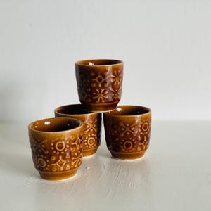 Op de afbeelding: Vier bruine keramische bekers met een herhalend patroon van cirkels, stippen en stervormige vormen. De bekers zijn in een piramidevorm gestapeld. De bekers zijn klein en lijken voor espresso of andere kleine drankjes te zijn.
