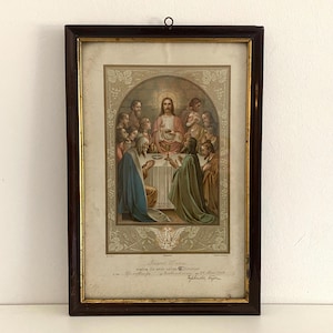 Op de afbeelding: Een ingelijst religieus drukwerk dat het Laatste Avondmaal voorstelt. De afbeelding toont Jezus Christus en zijn discipelen die rond een tafel zijn verzameld, waarbij Jezus een kelk vasthoudt. Het drukwerk is in een donkerhouten lijst met een gouden passe-partout.