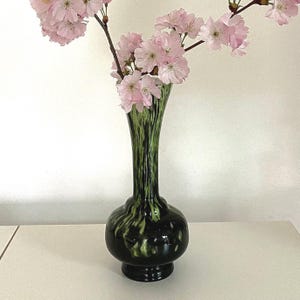 Può includere: Un vaso verde scuro con base rotonda e collo svasato, che contiene un ramo di fiori di ciliegio rosa. Il vaso presenta un motivo maculato e una finitura lucida. I fiori sono in piena fioritura, aggiungendo colore.