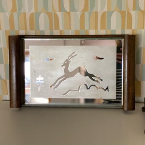 Op de afbeelding: Rechthoekige spiegel met een donkerbruin houten frame. De spiegel heeft een mat ontwerp van een springende gazelle, met gestileerde planten en wolken. De spiegel staat op een grijs oppervlak, met een behangachtergrond.
