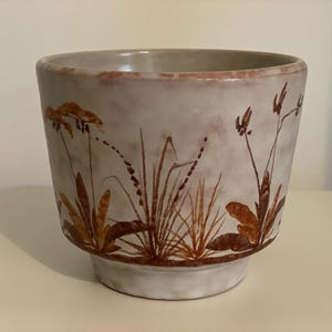 Könnte beinhalten: Ein weißer Keramiktopf mit braunem Blumendesign. Der Topf hat eine breite, runde Öffnung und einen leicht ausgestellten Boden.