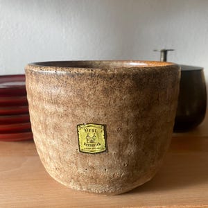 Könnte beinhalten: Ein brauner Keramiktopf mit strukturierter Oberfläche und einem kleinen Etikett mit der Aufschrift "Vest Keramik". Der Topf hat eine weite Öffnung und einen abgerundeten Boden. Weitere Keramikartikel sind im Hintergrund zu sehen.