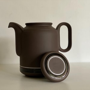 May include: A brown ceramic teapot with a matching lid. The teapot has a rounded body, a curved spout, and a large handle. The lid is off to the side, revealing a white interior rim. The teapot is a dark brown color.