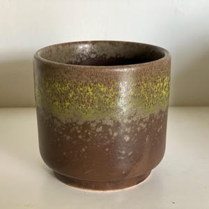 Könnte beinhalten: Eine Keramiktasse mit zylindrischer Form und leicht ausgestelltem Boden. Die Tasse weist einen Farbverlauf auf, der von einem dunkelbraunen Boden zu einem gesprenkelten grün-braunen Band am Rand übergeht. Der Innenraum ist dunkelbraun.