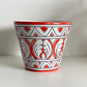 May include: A ceramic planter with a vibrant red rim and base. The white body is adorned with a repeating pattern of red and black geometric shapes and stylized leaf designs. The planter is suitable for small plants.