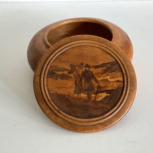 Könnte beinhalten: Ovale Schmuckschatulle aus hellem Holz mit Deckel. Der Deckel zeigt eine eingelegte Szene mit zwei Figuren in historischer Kleidung. Der Deckel ist dunkelbraun, was einen Kontrast zum hellen Holz der Schatulle bildet.