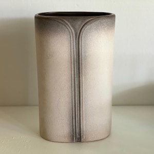 May include: A tall, cylindrical vase with a neutral color palette. The vase has a light beige body with a darker brown rim and a vertical, symmetrical design. The design is a series of parallel lines that run down the center of the vase.