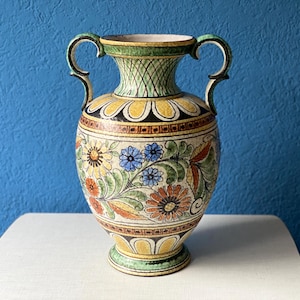 Può includere: Un vaso decorativo in ceramica con due manici, caratterizzato da un motivo floreale nei toni del giallo, blu, arancione e verde. Il vaso ha un corpo largo e un collo stretto, con un bordo verde e giallo. Il vaso è posto su una superficie bianca su sfondo blu.