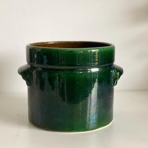Può includere: Vaso cilindrico in ceramica verde scuro con finitura lucida. Il vaso ha un'ampia apertura, interno marrone e due piccole maniglie arrotondate. Poggia su una superficie bianca.