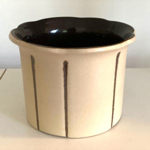 May include: A cream-coloured ceramic vase with a dark brown, scalloped rim. Vertical brown lines decorate the exterior. The interior is dark brown.