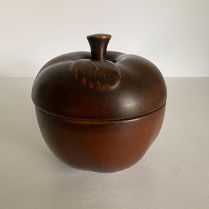 Pode incluir: Um recipiente de cerâmica castanha em forma de maçã com tampa. A tampa tem um caule e um detalhe de folha. O recipiente é de cor castanha quente e parece ser vidrado. O recipiente em forma de maçã é provavelmente para a cozinha ou decoração de casa.