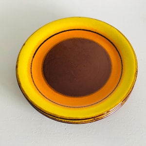 Könnte beinhalten: Ein Stapel von vier Keramikplatten mit Retro-Design. Jede Platte hat konzentrische Kreise in Gelb, Orange und Braun, mit einem dünnen schwarzen Strich, der die gelben und orangen Bänder trennt. Die Teller haben eine glänzende Oberfläche und einen leicht strukturierten Rand.