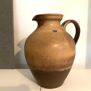 May include: A large, brown ceramic pitcher with a wide mouth and a handle. The pitcher has a mottled glaze with a darker brown band around the bottom.
