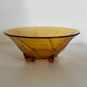 Op de afbeelding: Amberkleurige glazen kom met een zwarte rand en drie kleine voetjes. De kom heeft een breed, ondiep ontwerp met een licht getextureerd oppervlak. Het glas heeft een warme, gouden tint en de kom is waarschijnlijk vintage of retro.
