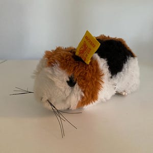 Op de afbeelding: Een klein, pluche speelgoed cavia met bruin, wit en zwart haar. Het speelgoed heeft een geel label met de tekst "G/N 2322" erop.